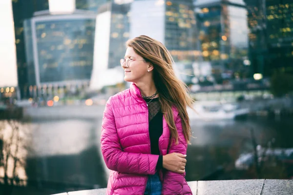 Young happy beautiful woman in glasses with long red hair in a bright pink jacket enjoying life in big city