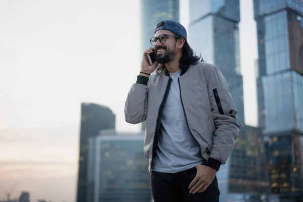Attractive brunette man in a cap with glasses in big city talking on a mobile phone