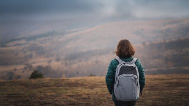 Gri sırt çantalı kadın gezgin soğuk ve bulutlu bir günde sırtını bir dağ manzarasına dayar.