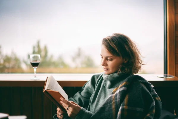 緑のセーターの若い女性は山の景色と彼女の国の家の窓の近くに本とワインを飲む読み取り — ストック写真