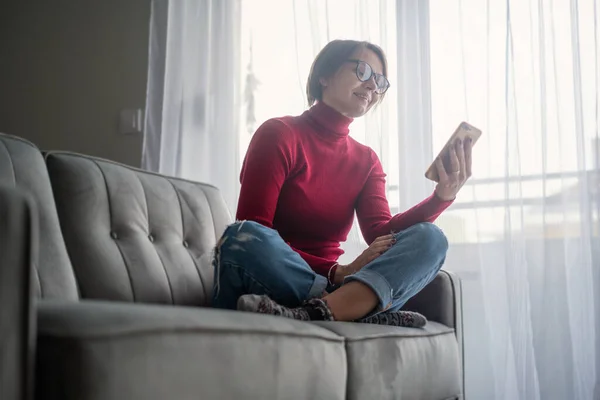 Mulher feliz sorridente bonita em uma gola alta vermelha em casa em um sofá cinza sentado com um telefone celular em suas mãos — Fotografia de Stock