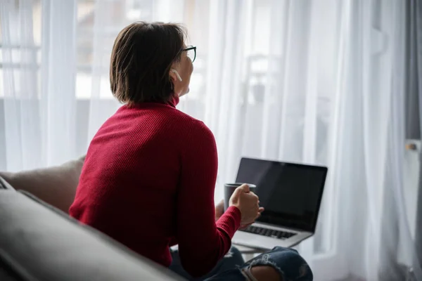 Красивая улыбающаяся счастливая женщина в очках в красной водолазке дома сидит дома на диване перед экраном ноутбука с кружкой кофе — стоковое фото