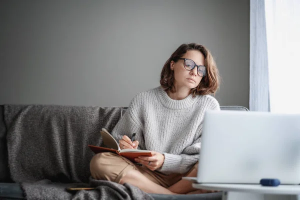 Belle jeune étudiante étudiant à la maison en ligne assis dans un pull chaud sur le canapé devant un ordinateur portable prenant des notes dans un carnet — Photo