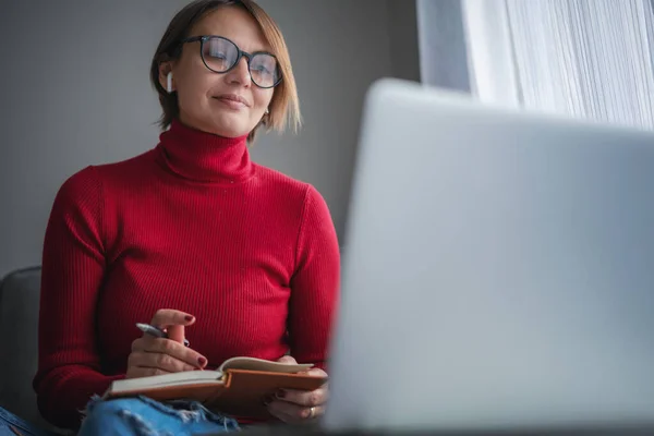 Hermosa mujer psicóloga adulta con gafas en casa sentada en casa en el sofá frente a la pantalla del ordenador portátil tomando notas durante la consulta de vídeo — Foto de Stock