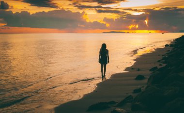 Tropik bir plajda, kumsalda yürüyen bir kızın silueti denizin üzerinde gün batımını seyrediyor.