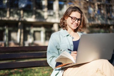 Parktaki bankta oturan ve güneşli bir günde dizüstü bilgisayarında yazan ciddi bir kız öğrenci..