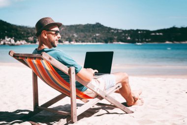 Kumsalda çalışan laptoplu çekici bir genç adam. Özgürlük, uzak çalışma, serbest çalışma, teknoloji, internet, seyahat