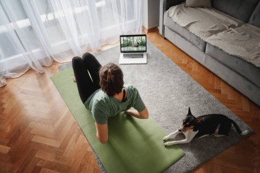 Neşeli, güzel bir kadın evde yoga yapıyor. Halıda yoga yapıyor. Yanında da köpeğiyle, spor ve akıl sağlığıyla yoga yapıyor.