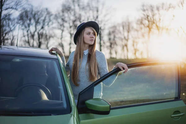 Vacker ung kvinna bär hatt reser med bil på landsbygden i bakgrunden av skog och solnedgång, lokala resor och äventyr koncept — Stockfoto