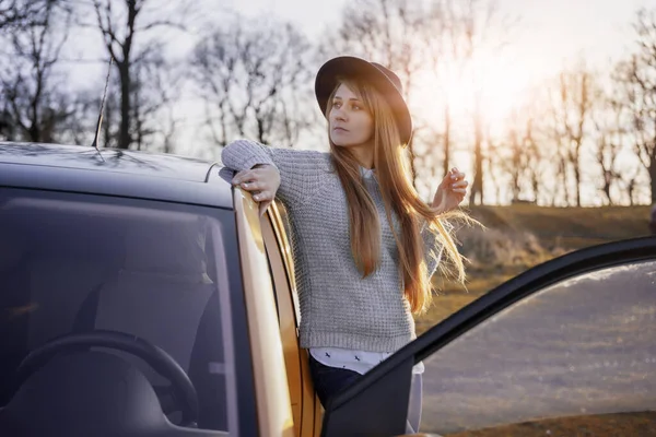 Schöne junge Frau mit Hut, die mit dem Auto in der Landschaft unterwegs ist, vor dem Hintergrund von Wald und Sonnenuntergang, lokalem Reise- und Abenteuerkonzept — Stockfoto