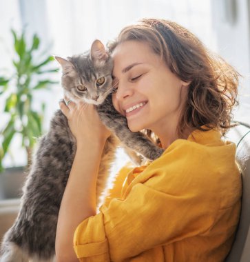 Evinde kollarında sevimli gri bir kediyle güzel, neşeli bir genç kadının yakın plan portresi.