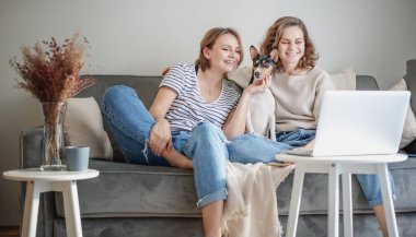 Güzel mutlu lezbiyen çift evdeki kanepede oturmuş evcil köpek Bassengi ile sohbet ediyorlar.