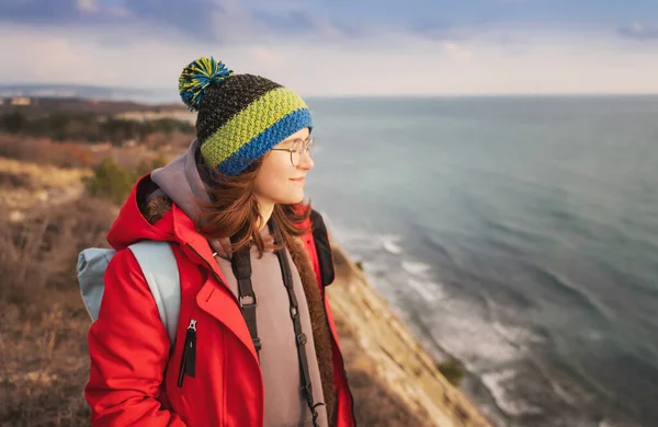 Young woman in cap and red jacket traveling enjoying the fresh air on the rocky seashore, solo travel
