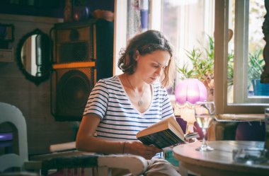 Kafede oturan ve elinde bir bardak şarapla kitap okuyan genç bir kadın.