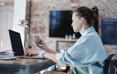 İnternette çalışan genç bir kadın ofisteki masada dizüstü bilgisayar ve akıllı telefon ekranına bakıyor, telefon ve defterler yakınlarda.