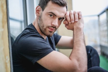 Şehre tepeden bakan balkonda siyah tişörtlü, sakallı yakışıklı, esmer adam.