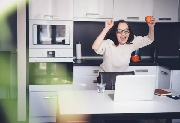 Счастливая Пожилая Женщина Апельсином Руках Стоит Дома Кухне Перед Экраном — стоковое фото