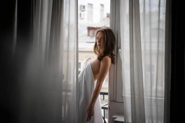 Piękna zmysłowa młoda kobieta z długimi włosami w białej koszuli stojąca przy oknie z zasłonami w mieszkaniu — Zdjęcie stockowe