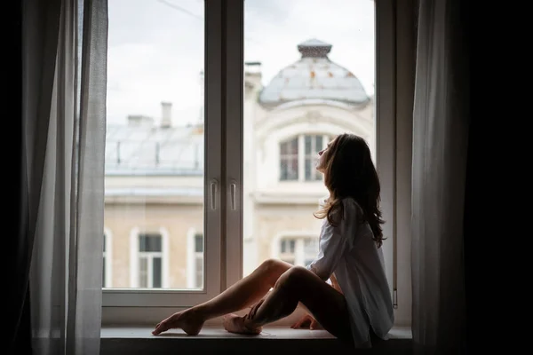 Bela sensual jovem com cabelos longos em uma camisa branca elegantemente sentado no peitoril da janela em um apartamento com vista para a cidade europeia — Fotografia de Stock