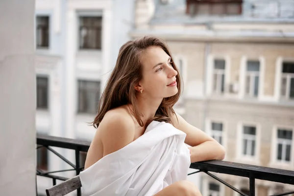 Красивая молодая чувственная женщина с длинными темными волосами, завернутыми в белую простыню, сидящая на балконе и смотрящая на город — стоковое фото
