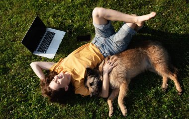 Güzel, mutlu, genç bir kadın dizüstü bilgisayarı ve köpeğiyle çimlerin üzerinde yatıyor.