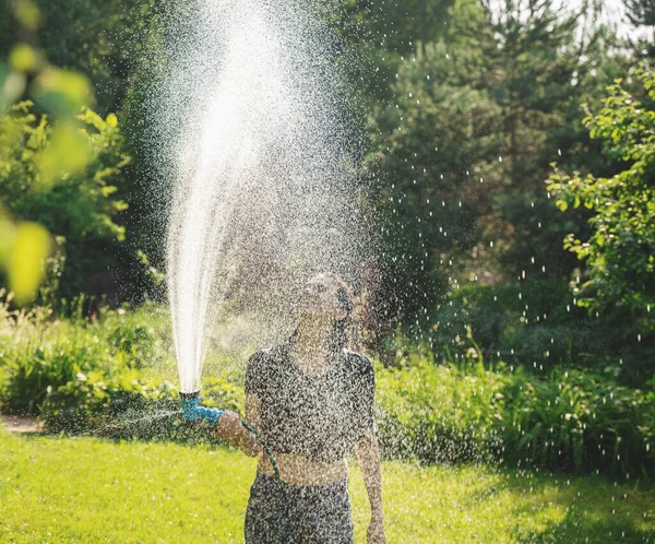美しい若い陽気な女性は暑い夏の日に緑の庭でスプリンクラーから水で身を注ぐ — ストック写真