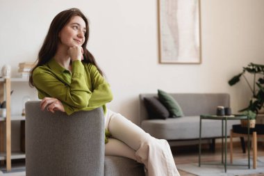 Yeşil bluzlu, genç, güzel bir kadın. Oturma odasındaki gri koltukta oturuyor.