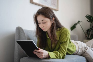 Evdeki kanepede tablet ve stil kullanarak uzanan yeşil bluzlu güzel, genç, mutlu esmer kadın..