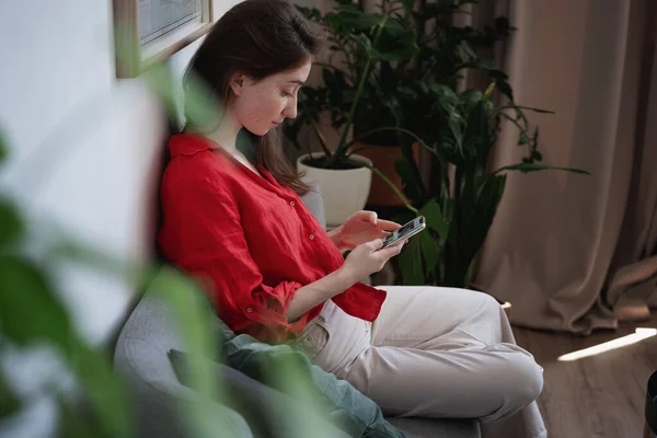 Piękna Młoda Elegancka Kobieta Czerwonej Bluzce Siedząca Kanapie Domu Smartfonem — Zdjęcie stockowe