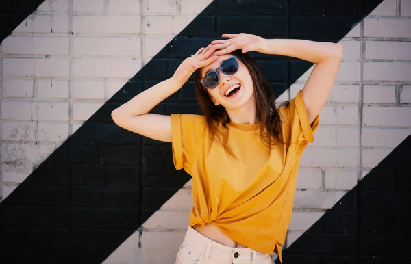 若い幸せな女の子の肖像画サングラスと黄色のTシャツ通りに壁の背景 — ストック写真