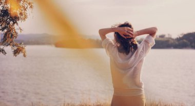 Güzel, genç, rahat, beyaz bluzlu bir kadın. Sırtı nehirde dururken doğanın tadını çıkarıyor.