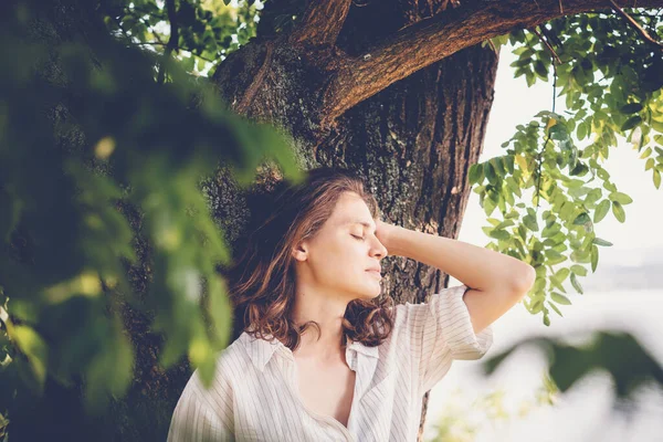 秋の日に木のそばに立ちながら自然を楽しむ白いブラウスの美しい若いリラックスした女性 — ストック写真