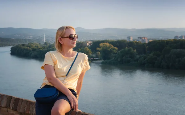 Beautiful adult blonde woman in sunglasses enjoying the view of the river at sunset — Stock Photo, Image
