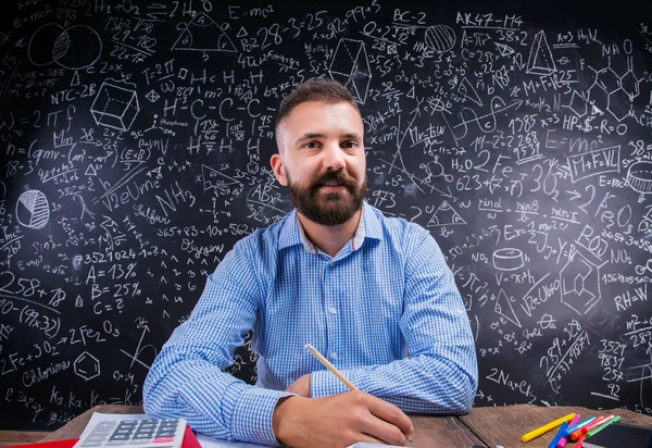 Happy teacher sitting at desk, school supplies, big blackboard — ストック写真