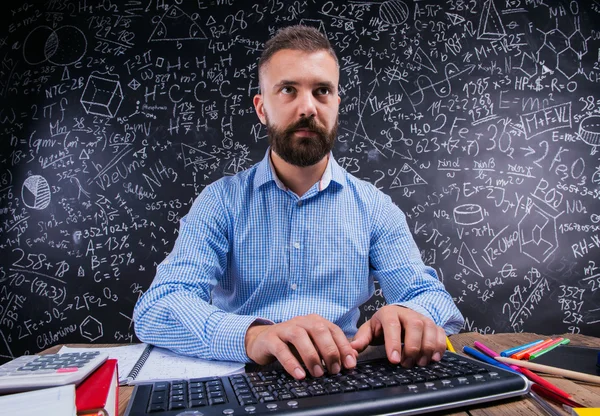 Happy teacher sitting at desk, school supplies, big blackboard — Φωτογραφία Αρχείου