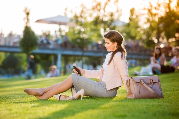 Mujer de negocios sentada en el parque revisando su teléfono inteligente, suma soleada —  Fotos de Stock