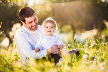Baba küçük oğlunu kucağına, çimenlerin üzerinde oturuyor.