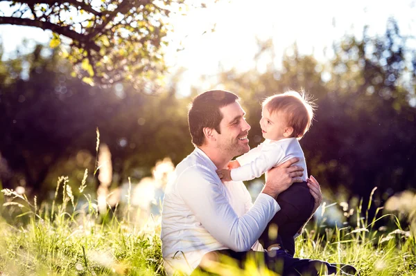 Baba küçük oğlunu kucağına, çimenlerin üzerinde oturuyor.