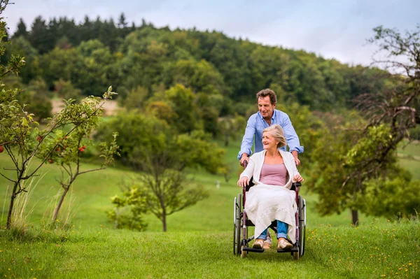 Ανώτερος άνθρωπος πιέζει γυναίκα το αναπηρικών αμαξιδίων, πράσινο φθινόπωρο φύση — Φωτογραφία Αρχείου