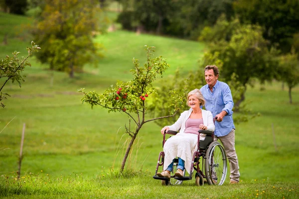 Homme âgé poussant femme en fauteuil roulant, vert automne nature — Photo