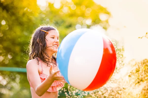 Mädchen im Bikini mit Ball am Sprinkler, Sommer — Stockfoto