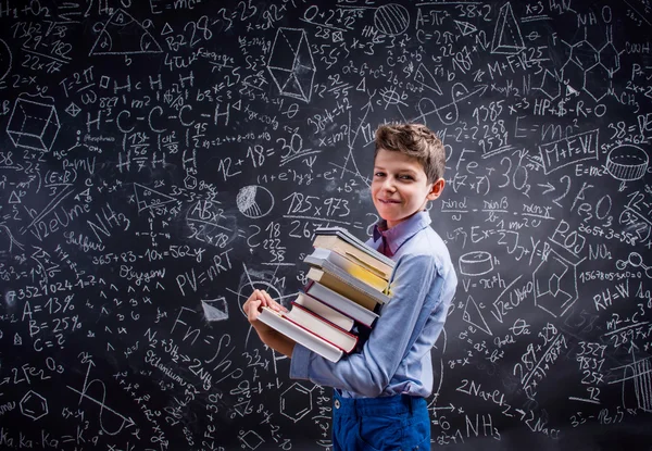 Boy with books against big blackboard with mathematical symbols — Stock Photo, Image