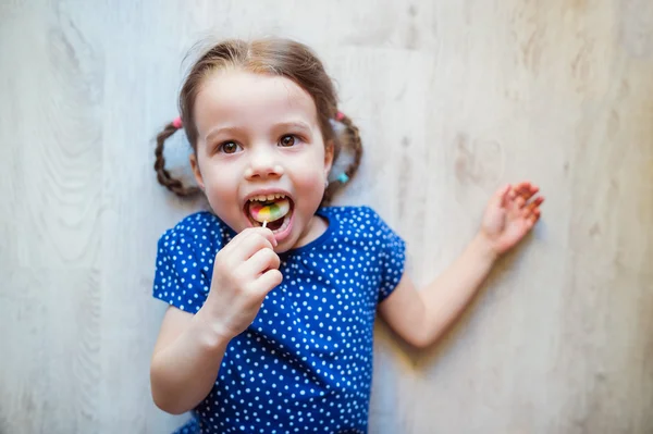 Niña tendida en el suelo, sonriendo, comiendo piruleta — Foto de Stock