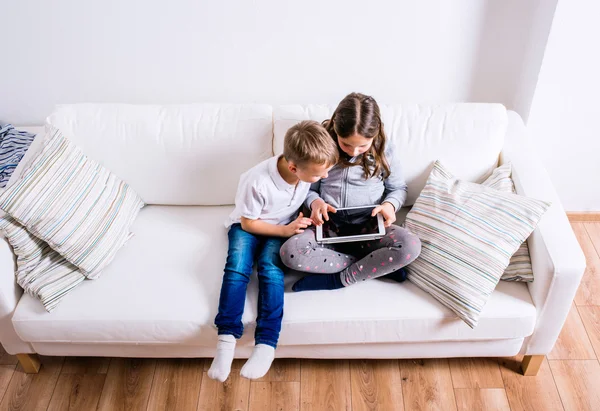 Anak-anak di rumah duduk di sofa, bermain dengan tablet — Stok Foto