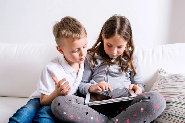 Anak-anak di rumah duduk di sofa, bermain dengan tablet — Stok Foto