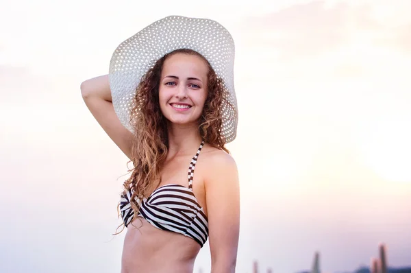 Mujer en la playa vistiendo bikini y sombrero, sonriendo, sosteniendo la cabeza —  Fotos de Stock