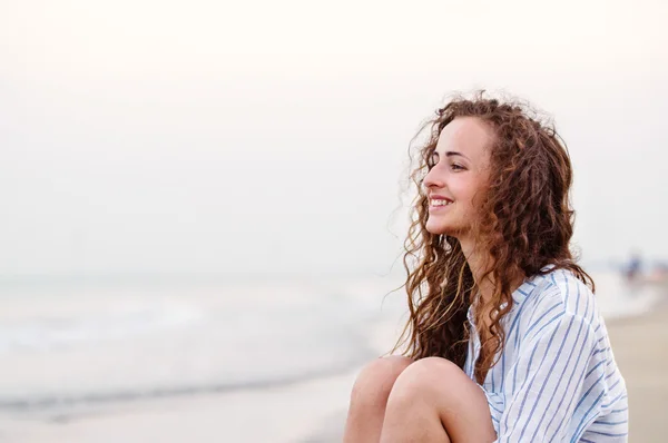 Piękna kobieta, siedząc na plaży w białej koszuli, uśmiechając się — Zdjęcie stockowe