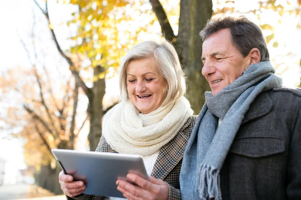 Senior couple with tablet sitting on bench. Autumn park. — Stock Photo, Image