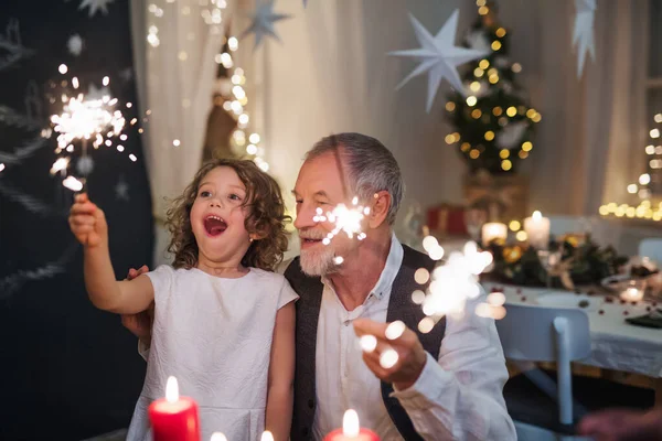 Senior grandfather with small granddaughter indoors at Christmas, sitting at table with sparklers. — Stock Photo, Image