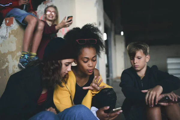 Grupa nastolatków gang siedzi wewnątrz w opuszczonym budynku, za pomocą smartfonów. — Zdjęcie stockowe